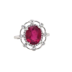 May include: A silver ring featuring a large, oval-cut ruby gemstone. The ruby is surrounded by a halo of smaller diamonds and a decorative outer ring of additional diamonds. The ring is set against a white background.