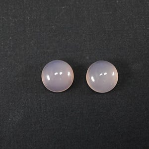 Puede incluir: Dos gemas redondas y pulidas, de color lavanda pálido. Las piedras lisas y reflectantes están sobre un fondo oscuro. Estos cabujones son adecuados para la fabricación de joyas u otros proyectos de artesanía.