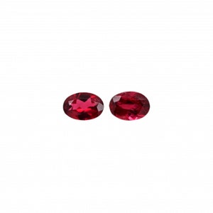 May include: Two oval-shaped, deep red gemstones. The gems are set against a white background. The stones have a polished, reflective surface, and are likely rubies or garnets. These gemstones are suitable for jewelry making.