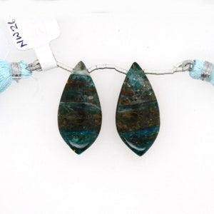 May include: Two teardrop-shaped gemstones with a mix of teal, brown, and gray colors. The stones are strung on a silver wire, with a light blue tassel on each side. The tag reads "NW26".