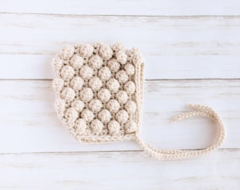 May include: A cream-colored crocheted baby bonnet with a textured bobble stitch pattern and a long tie.
