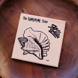 May include: A pink ceramic tile with a black and white illustration of a conch shell and the text "The Sunshine State Florida".