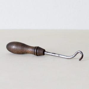 May include: An antique hook tool with a dark wooden handle and a curved metal hook. The handle is a dark brown color, and the metal hook is silver. The tool is designed for pulling or hooking.