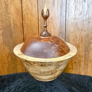 May include: A handcrafted wooden bowl with a lid and a decorative finial. The bowl is made of light and dark brown wood, with a smooth, polished finish. The lid is a darker shade of brown, and the finial is shaped like a flame.
