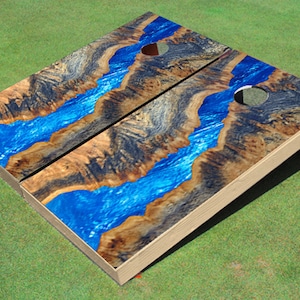 May include: A wooden cornhole game with a blue and brown river design. The boards are made of wood and have a natural finish. The holes are cut out of the boards and are painted black.