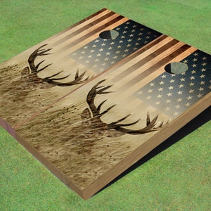 May include: A set of two wooden cornhole boards with a distressed American flag design and a silhouette of a deer's head in the background.
