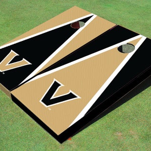 May include: A set of two black and gold cornhole boards with a white V logo. The boards have a black background with a gold triangle and a white stripe.
