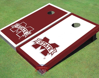 Può includere: Un set di due tavole da cornhole di dimensioni regolamentari con un design bianco e marrone. Le tavole presentano il logo della Mississippi State University con le parole "State" in uno stendardo sopra la lettera "M".