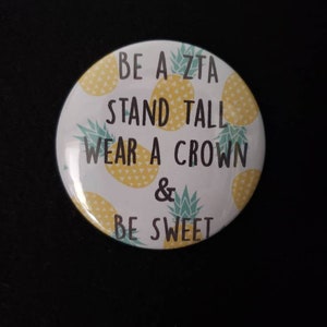May include: White button with a pineapple pattern and the text "Be a Zeta Stand Tall Wear a Crown & Be Sweet".