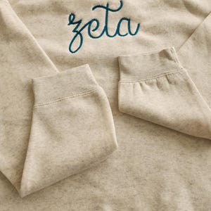 May include: A light beige sweatshirt with a teal blue embroidered word "zeta" on the front.