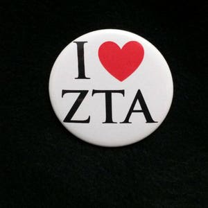 May include: A white button with the text "I ❤️ ZTA" in black letters and a red heart symbol. The button is round and set against a black background.