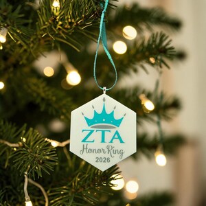 May include: A hexagonal white ornament hanging from a teal ribbon. The ornament features a teal crown graphic above the text ZTA, the words Honor Ring in a cursive script, and the year 2026 printed in a dark gray font on the front surface.