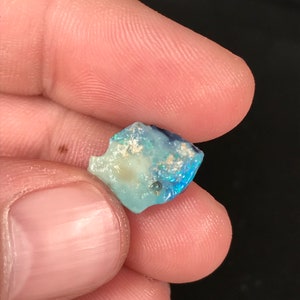 May include: A small, rough-cut opal gemstone with a mix of blue, white, and iridescent colors. The stone is held between fingers, showcasing its natural, irregular shape and subtle color variations. The opal has a polished, reflective surface.