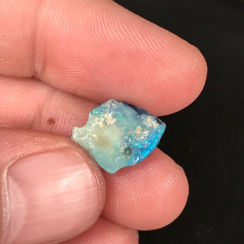 May include: A small, rough-cut opal gemstone with a mix of blue, white, and iridescent colors. The stone is held between fingers, showcasing its natural, irregular shape and subtle color variations. The opal has a polished, reflective surface.
