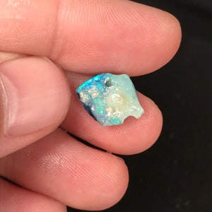 May include: A small, irregularly shaped opal gemstone with a mix of blue, green, and white colors. The opal is held between fingers, showcasing its natural, raw form. The stone has a rough texture and a small hole.