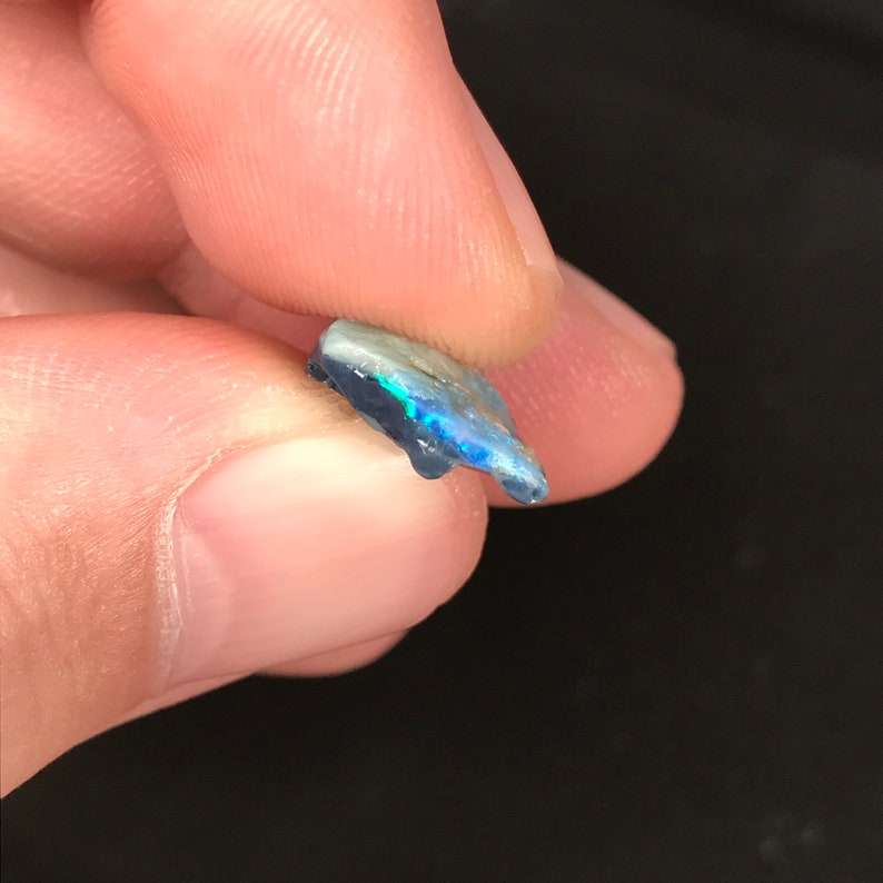 May include: A small, irregularly shaped opal gemstone with a dark blue base and iridescent flashes of green and blue. The opal is held between two fingers, showcasing its natural, rough texture and vibrant play of color.