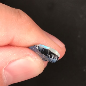 May include: A small, rough-cut opal gemstone held between fingers. The opal displays a mix of colors, including blue, black, and white, with a shimmering, iridescent quality. The stone has a jagged edge and a natural, unpolished appearance.