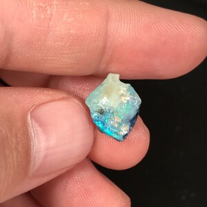 May include: A small, rough-cut opal gemstone with a mix of colors. The opal displays a play of color with flashes of blue, green, and hints of yellow and white. The gemstone is held between two fingers.