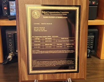 May include: A brown wooden plaque with a gold border and a black background. The plaque has the text "Federal Communications Commission Wireless Telecommunications Bureau RADIO STATION AUTHORIZATION" at the top. The plaque also has the text "LICENSEE: SMITH CRAIG B SMITH, CRAIG B 892 SKYLINE DR FORSYTH, MO 65653" in the middle. The plaque also has the text "FCC Registration Number (FRN): 0020346664 Call Sign WRKG680 File Number 0009350042 Grant Date 01-01-2021 Effective Date 01-01-2021 Radio Service ZA-General Mobile Radio (GMS) Prist Date 01-01-2021 Expiration Date 01-01-2031" at the bottom. The plaque is on a clear acrylic stand.