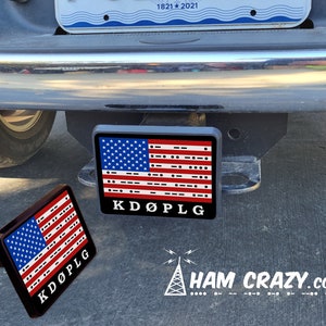 May include: A black and red American flag with the letters "KDOPLG" printed below it. The flag is made of a material that looks like plastic and is attached to a black metal hitch cover. The hitch cover is attached to a black truck. The text "HAM CRAZY.COM" is printed below the flag with a white antenna symbol.