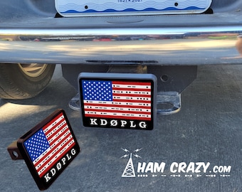 May include: A black and red American flag with the letters "KDOPLG" printed below it. The flag is made of a material that looks like plastic and is attached to a black metal hitch cover. The hitch cover is attached to a black truck. The text "HAM CRAZY.COM" is printed below the flag with a white antenna symbol.