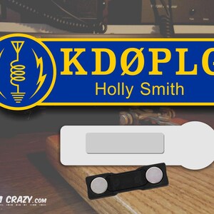 May include: A blue and yellow name tag with the call sign "KD&Oslash;PLG" and the name "Holly Smith" printed on it. The name tag is attached to a magnetic backing.