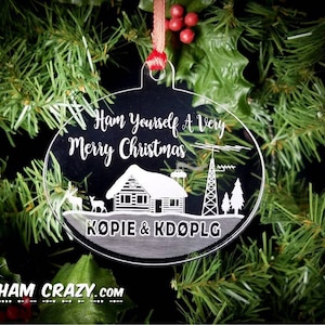 May include: A clear glass Christmas bauble with the text "Ham Yourself A Very Merry Christmas" and "KOPIE & KDOPLG" etched on it. The bauble also features a small cabin, two deer, and a radio antenna.