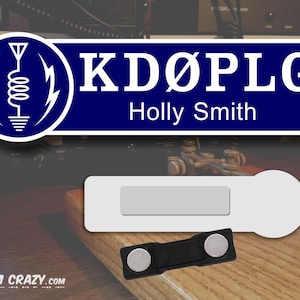May include: A blue and white name tag with the call sign "KD&Oslash;PLG" and the name "Holly Smith" printed on it. The tag has a white background with a blue border and a white antenna symbol on the left side. The tag is attached to a black magnetic clip.