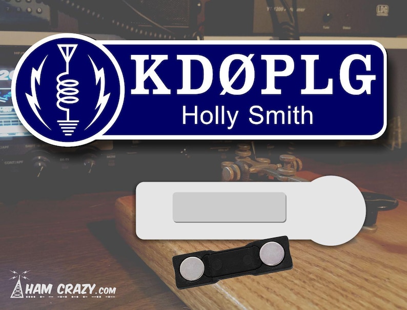 May include: A blue and white name tag with the call sign "KD&Oslash;PLG" and the name "Holly Smith" printed on it. The tag has a white background with a blue border and a white antenna symbol on the left side. The tag is attached to a black magnetic clip.