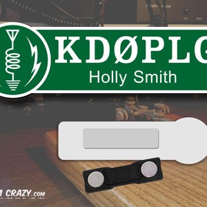 May include: Green and white sign with the call sign "KD&Oslash;PLG" and the name "Holly Smith" below. The sign has a green circle with a white antenna and lightning bolts on the left side. A white name tag with a black magnetic backing.