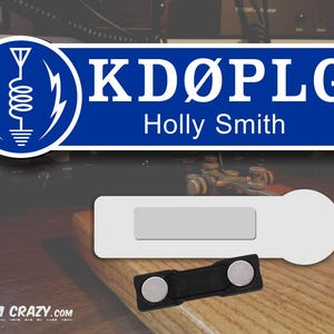 May include: A blue and white name tag with the call sign "KD&Oslash;PLG" and the name "Holly Smith". The tag has a white background with a blue border and a white antenna symbol in the top left corner. The tag is attached to a black magnetic clip.