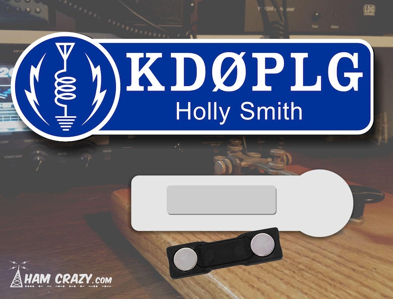 May include: A blue and white name tag with the call sign "KD&Oslash;PLG" and the name "Holly Smith". The tag has a white background with a blue border and a white antenna symbol in the top left corner. The tag is attached to a black magnetic clip.