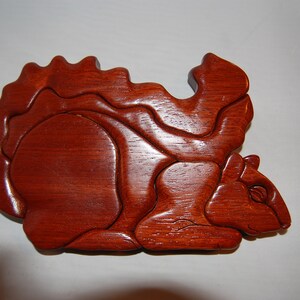 May include: A wooden squirrel sculpture made of multiple interlocking pieces. The sculpture is a reddish-brown color and has a detailed, realistic design.