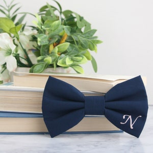 May include: A navy blue bow tie with a pink embroidered letter "N" on a stack of books.