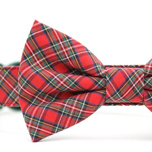 May include: A red plaid bow tie attached to a matching fabric collar. The fabric features a classic tartan pattern with intersecting lines in shades of green, yellow, white, and dark blue against a vibrant red background, creating a structured bow.
