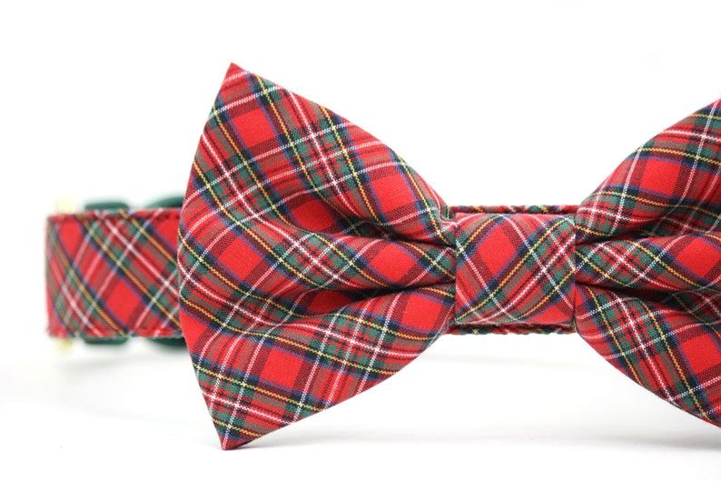 May include: A red plaid bow tie attached to a matching fabric collar. The fabric features a classic tartan pattern with intersecting lines in shades of green, yellow, white, and dark blue against a vibrant red background, creating a structured bow.