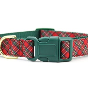 May include: A pet collar featuring a red, green, and yellow plaid fabric pattern. The collar includes a dark green plastic side-release buckle and a gold-toned metal D-ring for attachment. The inner lining of the collar is a solid dark green.