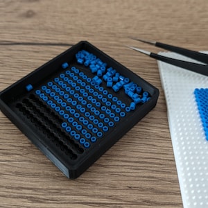 May include: A black plastic tray with a grid pattern filled with blue plastic beads. A white pegboard with a row of blue beads is in the background.