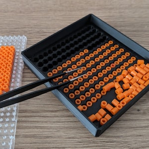 May include: A black tray with orange plastic beads, some of which are being picked up with tweezers. The tray is divided into sections, and there are many beads in each section.