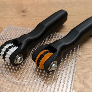 Puede incluir: Dos herramientas con mango negro y ruedas de colores diferentes. Las ruedas son de plástico y tienen una superficie texturizada. Las herramientas están sobre una superficie de plástico transparente con un patrón de cuadrícula.