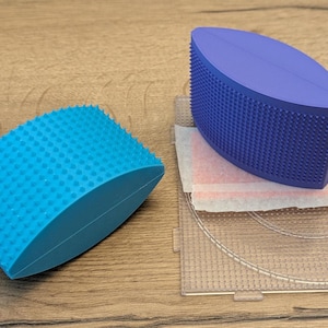 May include: Two curved, textured massage tools in the image. One is bright blue, the other is a deep purple. The tools have a series of small, rounded bumps on the curved surface. The tools are on a wooden surface.