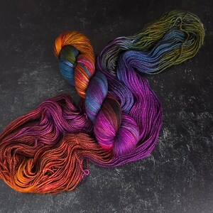 Puede incluir: Una colecci&oacute;n de ovillos de lana de colores en tonos de morado, naranja, verde y azul. La lana est&aacute; dispuesta en un patr&oacute;n suelto y fluido.