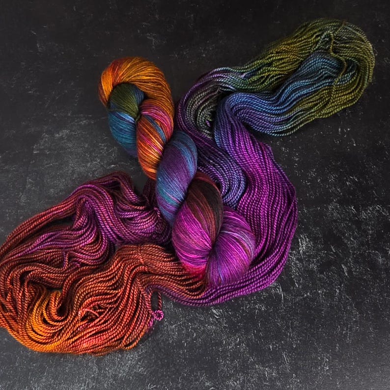 Puede incluir: Una colecci&oacute;n de ovillos de lana de colores en tonos de morado, naranja, verde y azul. La lana est&aacute; dispuesta en un patr&oacute;n suelto y fluido.