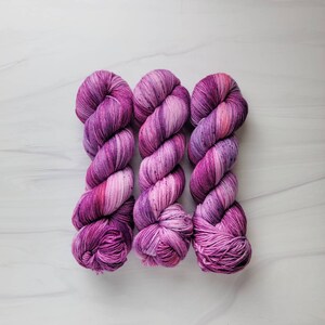 May include: Three skeins of yarn in shades of purple and pink. The yarn is twisted and has a variegated color pattern.