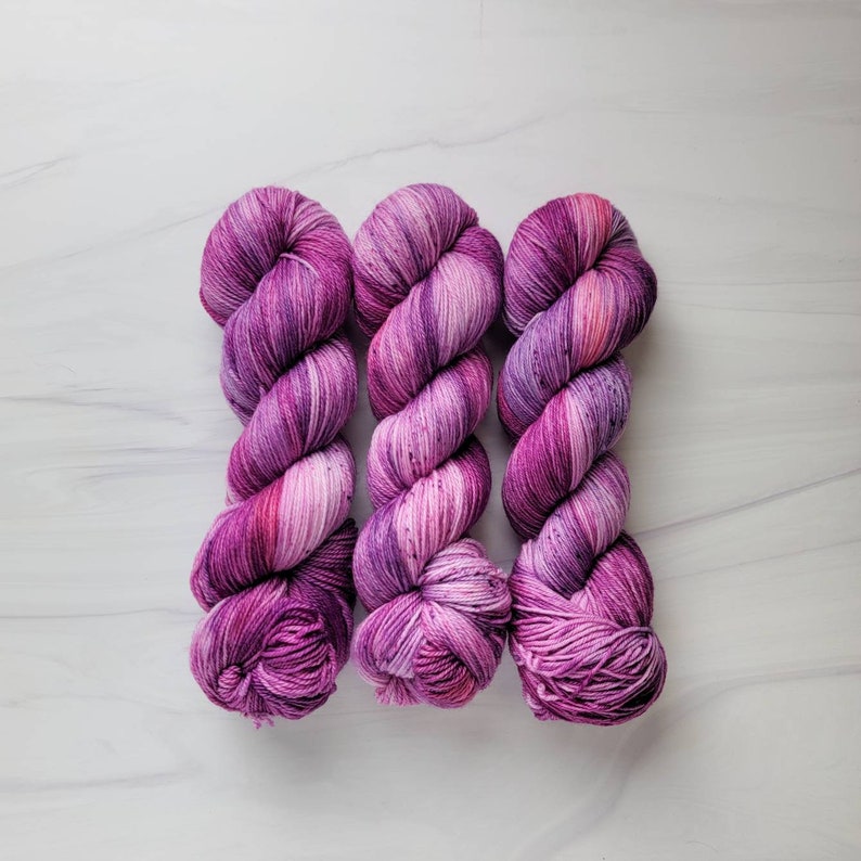 May include: Three skeins of yarn in shades of purple and pink. The yarn is twisted and has a variegated color pattern.
