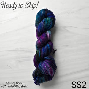 May include: A skein of Squishy Sock yarn in shades of blue, purple, and teal. The yarn is labeled "SS2" and includes the text "437 yards/100g skein". The words "Ready to Ship!" are also visible.
