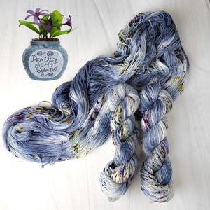 K&ouml;nnte beinhalten: Handgef&auml;rbtes Garn in Blau-, Wei&szlig;- und Sprenkel-T&ouml;nen mit Lila, Gelb und Gr&uuml;n. Das Garn wird mit einer kleinen blauen Vase mit dem Text "Deadly Night Shade" und lila Blumen pr&auml;sentiert.
