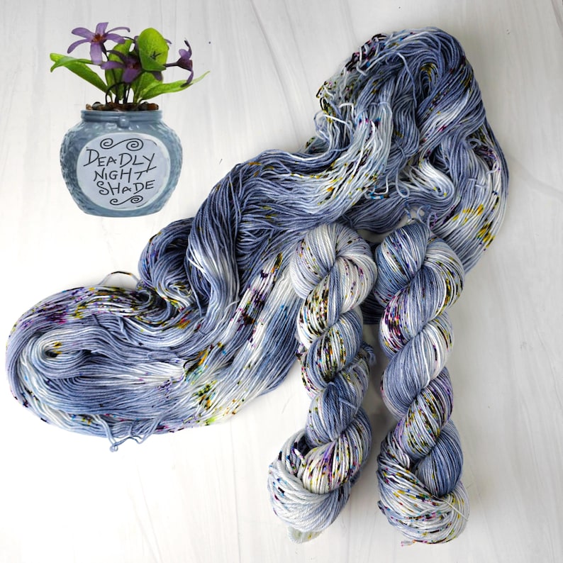 K&ouml;nnte beinhalten: Handgef&auml;rbtes Garn in Blau-, Wei&szlig;- und Sprenkel-T&ouml;nen mit Lila, Gelb und Gr&uuml;n. Das Garn wird mit einer kleinen blauen Vase mit dem Text "Deadly Night Shade" und lila Blumen pr&auml;sentiert.