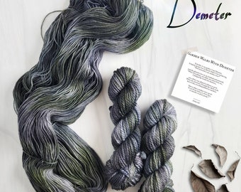Può includere: Due gomitoli di filato in tonalità di verde, grigio e viola. Il filato è disposto in un motivo sciolto e fluido. Il filato è etichettato "Demeter: Garden Walks with Demeter".