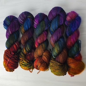 Puede incluir: Cinco ovillos de hilo de colores en tonos de morado, azul, verde, naranja y rojo. El hilo est&aacute; dispuesto en una fila, con los extremos de cada ovillo superpuestos.